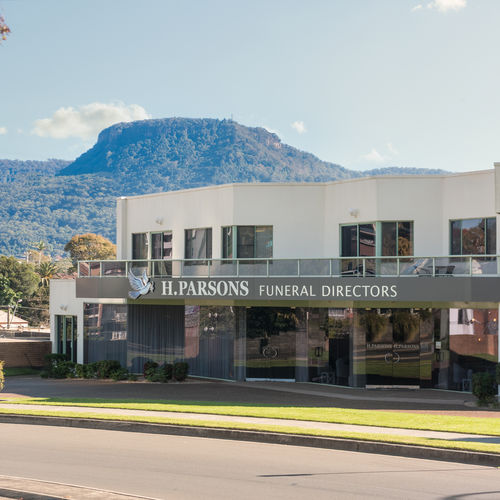 A modern white building with "H. Parsons Funeral Directors" signage on the front, set against a backdrop of a tree-covered mountain under a clear sky. by H Parsons Funeral Directors