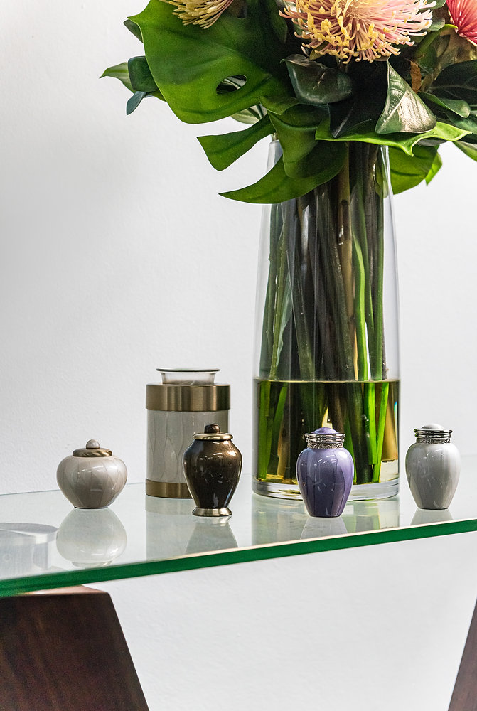 Four small decorative urns and a larger container are displayed on a glass table. Behind them is a tall glass vase with water, green leaves, and large, pinkish flowers—a serene setting often chosen for affordable funerals or direct cremation services. by H Parsons Funeral Directors