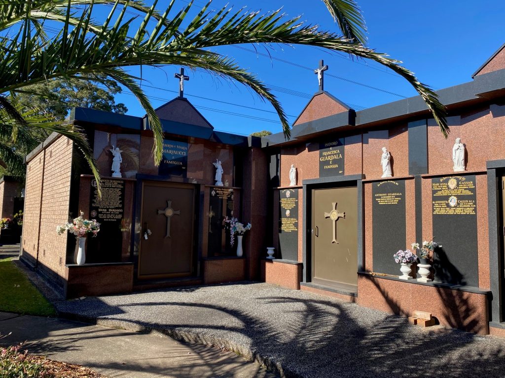 A mausoleum with brown and black stone walls, gold crosses on the doors, flower arrangements, and white statues of religious figures above inscriptions—palm fronds arch in the foreground under a bright blue sky, reminding visitors of NSW funeral claims processes. by H Parsons Funeral Directors
