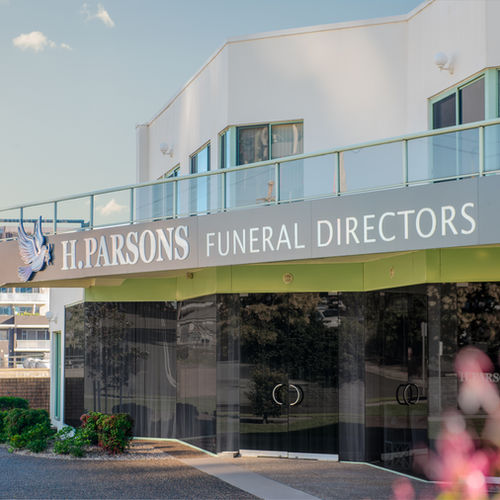 A modern building with a sign reading "H. Parsons Funeral Directors" above glass doors, with a clear sky and greenery reflected in the windows. by H Parsons Funeral Directors