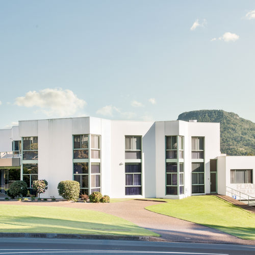 A modern, white two-storey building with large windows sits on a neatly landscaped lawn. A rounded hill rises in the background under a clear blue sky with a few clouds. by H Parsons Funeral Directors
