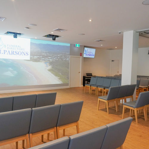 A modern funeral chapel with rows of grey chairs facing a large screen displaying a beach scene and the text "Funeral Directors T. Parsons." The space has wooden floors and white walls. by H Parsons Funeral Directors
