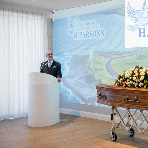 A man in a suit stands at a white lectern, speaking at a funeral service. A coffin adorned with white flowers is nearby, and a projection with "Funeral Directors H. Parsons" is displayed in the background. by H Parsons Funeral Directors