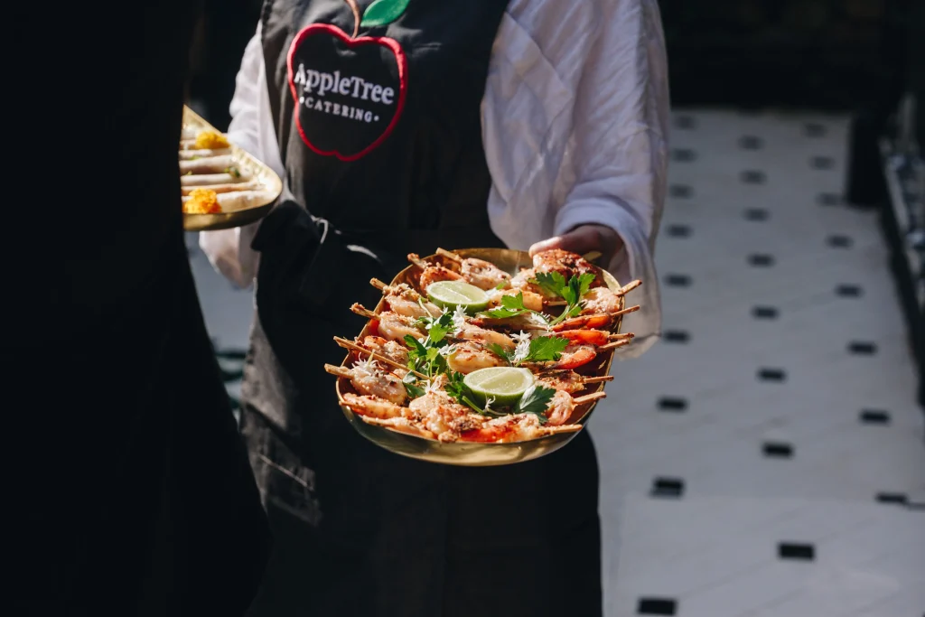 A waiter wearing a black "AppleTree Catering" apron holds a platter of skewered prawns garnished with lime wedges and herbs, showcasing AppleTree’s commitment to sustainable catering. Another plate of starters is partially visible in the background. by H Parsons Funeral Directors