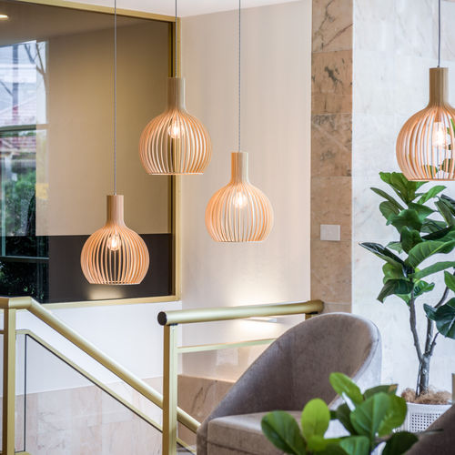 A modern interior with three wooden pendant lights, a potted plant, a grey chair, and gold railings. The space features marble and light-coloured walls, large windows, and a contemporary, cosy atmosphere. by H Parsons Funeral Directors