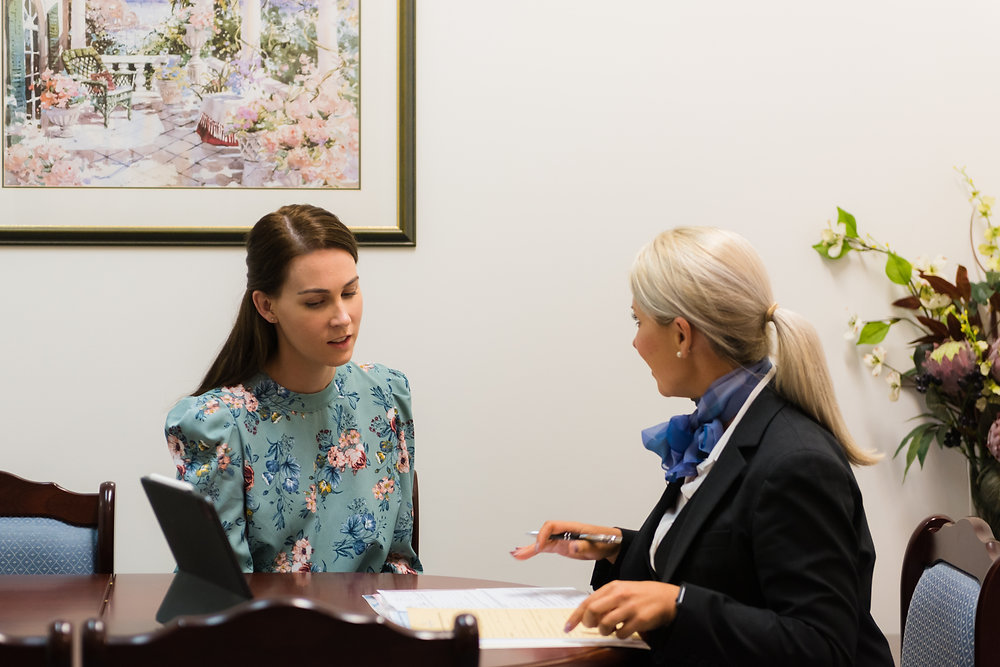 Two women sit at a table discussing paperwork. One in a floral dress listens attentively, while the other, dressed in a suit with a blue scarf, gestures with a pen as they review funeral insurance cost. A laptop and documents are on the table; flowers and art decorate the room. by H Parsons Funeral Directors