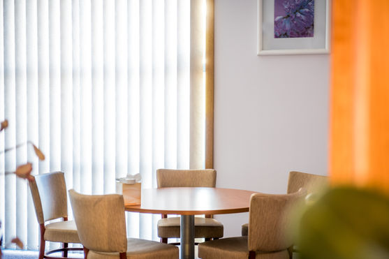 A round wooden table surrounded by four beige upholstered chairs sits by a large window with vertical blinds; a box of tissues is on the table and a framed picture hangs on the white wall. by H Parsons Funeral Directors