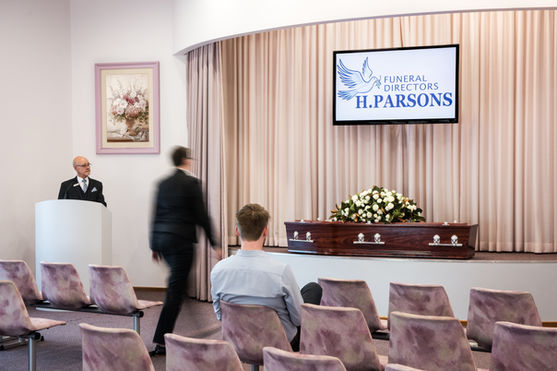 A funeral service room with rows of empty chairs, a man seated, another walking by, and a man standing at a lectern. A coffin with white flowers is in front of closed curtains, and a screen displays "Funeral Directors H. Parsons. by H Parsons Funeral Directors