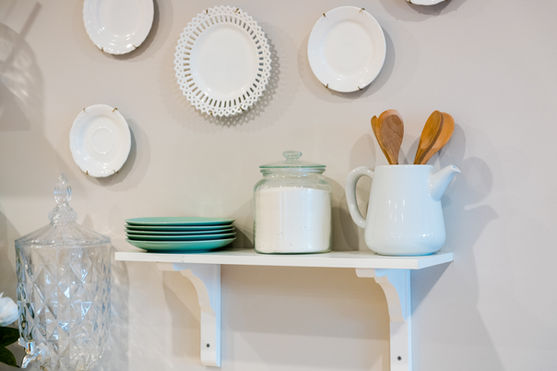 A white shelf holds green plates, a glass jar of sugar, and a white jug with wooden utensils. Decorative white plates are arranged on the wall above the shelf. by H Parsons Funeral Directors