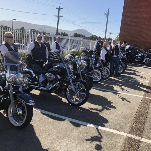 A group of people wearing waistcoats and sunglasses stand beside a row of parked motorbikes in a sunny outdoor car park, gathering in support of a personalised funeral service, with mountains and buildings visible beyond the fence. by H Parsons Funeral Directors
