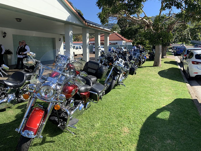 A row of shiny motorbikes, including a unique motorbike hearse, is parked on a grassy lawn beside a house, with people gathered under a porch in the background. Trees and parked cars line the street under a bright, sunny sky. by H Parsons Funeral Directors
