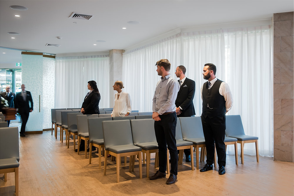 Five people stand spaced apart in a modern, sunlit room with rows of grey chairs and large white curtains, reflecting a quiet moment often seen when arranging a funeral or considering a funeral planning checklist. by H Parsons Funeral Directors