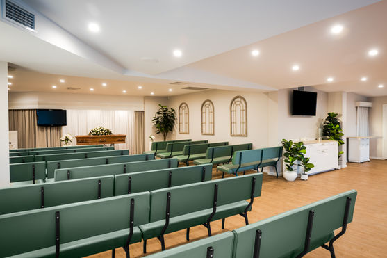 A modern funeral parlour chapel with green cushioned benches, potted plants, wooden flooring, and a coffin adorned with white flowers at the front. The room is well-lit with recessed ceiling lights. by H Parsons Funeral Directors