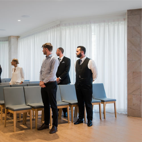 Four people, three men in formal attire and one woman in a white blouse, stand and sit among rows of empty grey chairs in a bright room with large windows and sheer white curtains. by H Parsons Funeral Directors