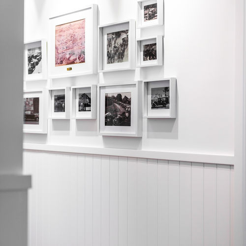 A white wall displays a collage of framed black-and-white photos and artwork, arranged in a grid pattern above white panelling. The photo is taken from the side, partially framed by a white doorway. by H Parsons Funeral Directors