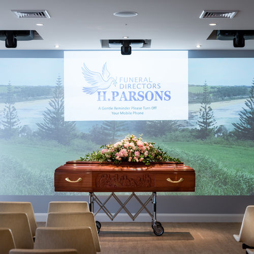 A wooden coffin with a flower arrangement rests at the front of a funeral service room. Behind it is a screen displaying "FUNERAL DIRECTORS H. PARSONS" and a reminder to switch off mobile phones. Empty chairs are visible. by H Parsons Funeral Directors