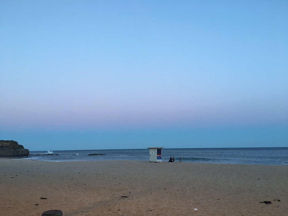A sandy beach at dusk with a lifeguard hut near the shoreline. Two people sit by the water, perhaps sharing condolence phrases as the calm sea meets a soft blue and pink sky. A rocky cliff rises quietly on the left. by H Parsons Funeral Directors