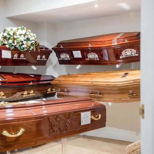 A display room with several polished wooden coffins, some adorned with decorative handles and panels. A bouquet of white flowers rests on one coffin. The room is well-lit and tidy. by H Parsons Funeral Directors