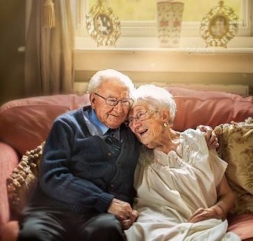 An elderly couple sit close together on a sofa, smiling and embracing lovingly. Soft sunlight filters through the window behind them, creating a warm, cosy atmosphere—perfect for heartfelt funeral tributes and memorial announcements. by H Parsons Funeral Directors