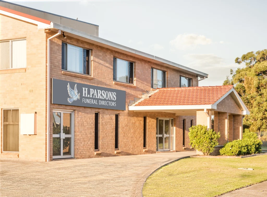 A two-storey brick building with a sign reading "H. Parsons Funeral Directors" above the entrance, this local funeral home is surrounded by a paved driveway and some greenery on a sunny day. by H Parsons Funeral Directors