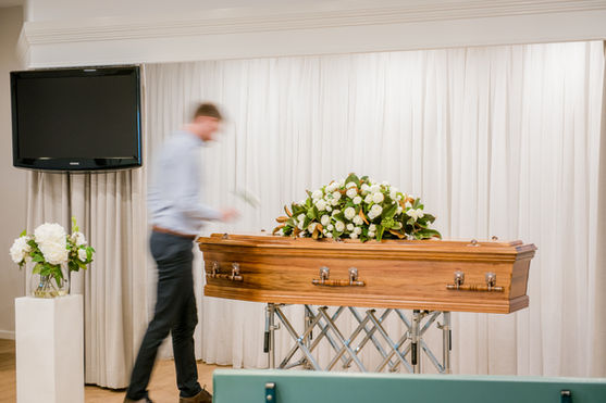 A wooden coffin with a large white flower arrangement on top is displayed in a room with white curtains. A blurred person stands beside it. A vase of white flowers is on a pedestal, and a TV is mounted on the wall. by H Parsons Funeral Directors