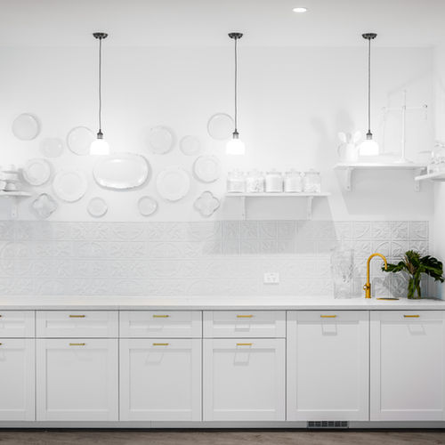 Modern white kitchen with minimalist design, featuring white cupboards, a gold tap, pendant lights, floating shelves with jars, and decorative white plates on the wall above the splashback. by H Parsons Funeral Directors