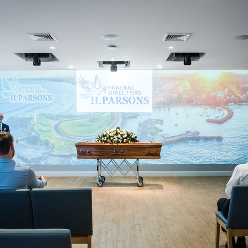 A coffin adorned with white and yellow flowers is displayed at the front of a modern funeral chapel, with “Funeral Directors H. Parsons” and a coastal landscape projected on the wall behind it. by H Parsons Funeral Directors