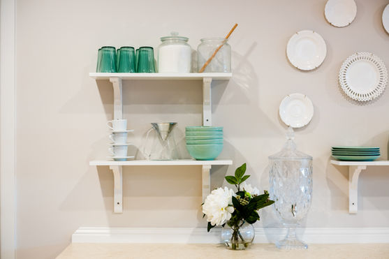 Two white wall shelves display green-tinted glasses, stacked bowls, plates, jars, and cups. A clear glass jar with a lid and a vase with white flowers sit below. Decorative plates hang on the beige wall. by H Parsons Funeral Directors