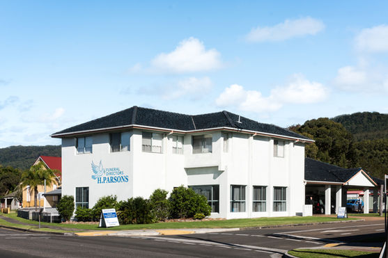 A two-storey white building on a street corner has a sign reading "Funeral Directors H. Parsons" and is surrounded by greenery, with hills and a partly cloudy sky in the background. by H Parsons Funeral Directors