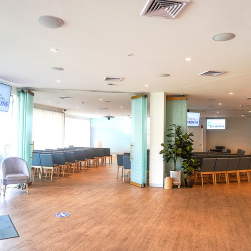 A spacious, modern room with rows of grey cushioned chairs, wooden flooring, potted plants, and large windows allowing natural light. Two screens are visible at the far end, and glass doors are partially open. by H Parsons Funeral Directors