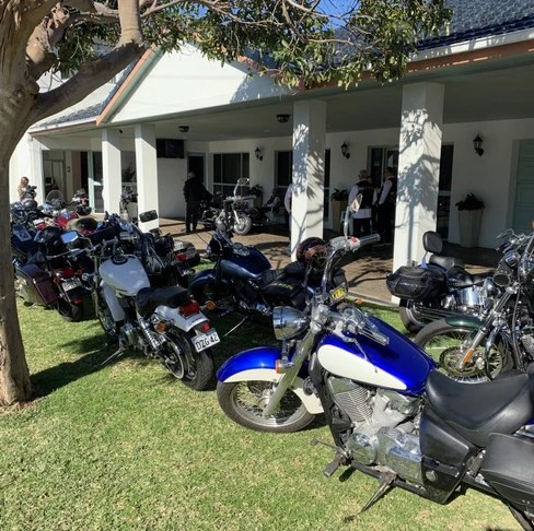 Several motorbikes are parked on grass outside a white building with a covered veranda, where people gather under the shade of a tree—a fitting scene for a personalised biker funeral on a sunny day. by H Parsons Funeral Directors