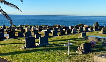 A cemetery with rows of dark headstones and a white cross overlooks the sea on a sunny day; a palm tree is on the left, and colourful flowers are placed by some graves. by H Parsons Funeral Directors