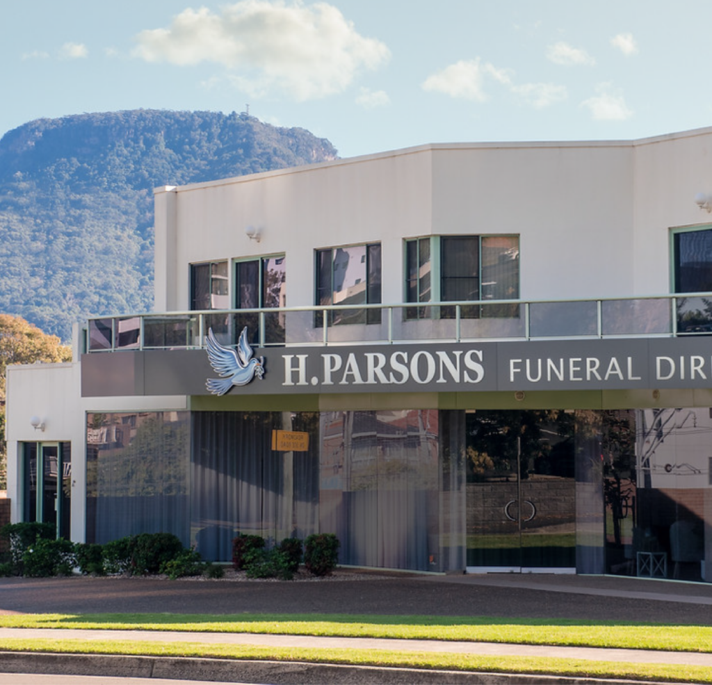 A modern white building with large windows and signage reading "H. Parsons Funeral Directors," surrounded by greenery, with a mountain and blue sky in the background. by H Parsons Funeral Directors