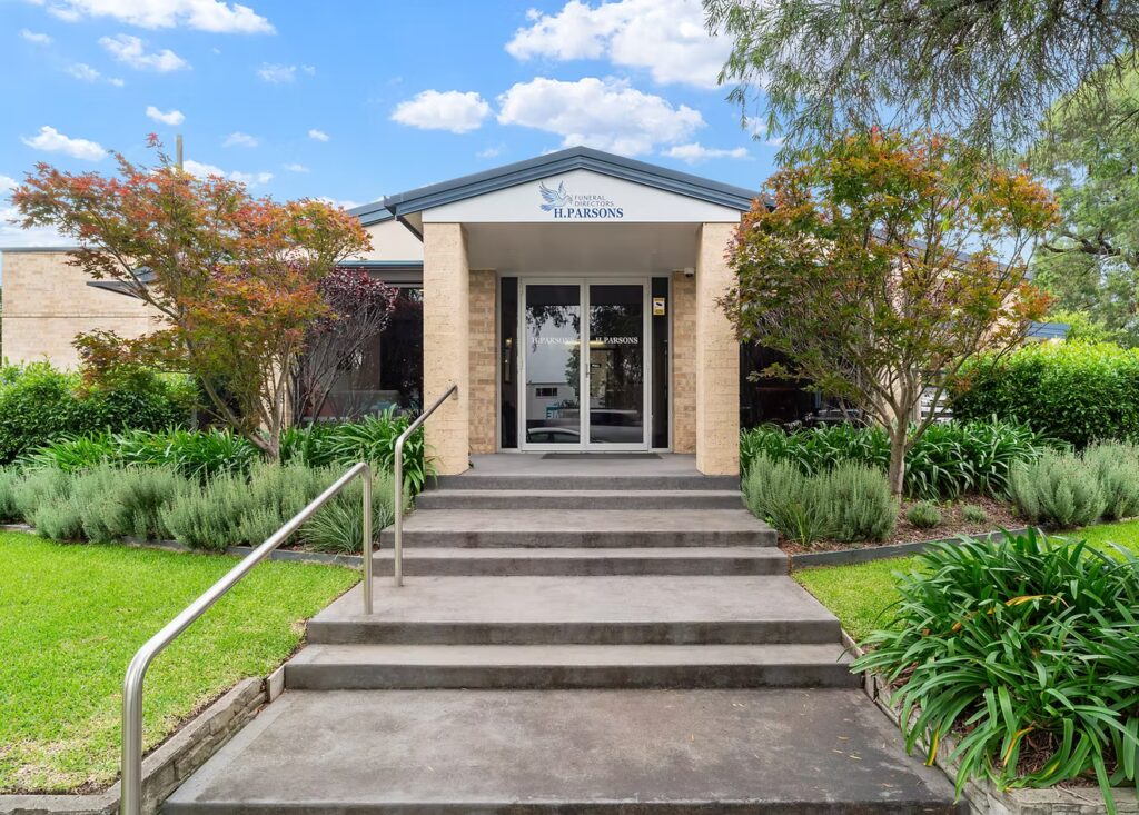Single-storey building entrance with a grey ramp and steps, metal handrails, glass double doors, and gardens with green shrubs and small trees on either side. Blue sky with scattered clouds above. by H Parsons Funeral Directors