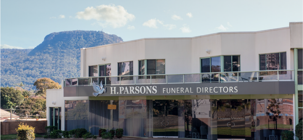 A modern white building with large windows and a sign reading "H. Parsons Funeral Directors", set against a backdrop of a mountain and clear blue sky. by H Parsons Funeral Directors