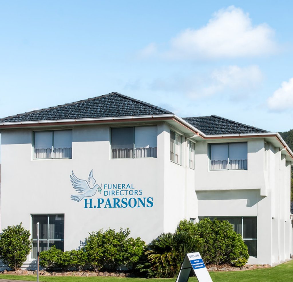 A white two-storey building with “Funeral Directors H. Parsons” and a bird logo on the wall, surrounded by green shrubs under a blue sky. by H Parsons Funeral Directors