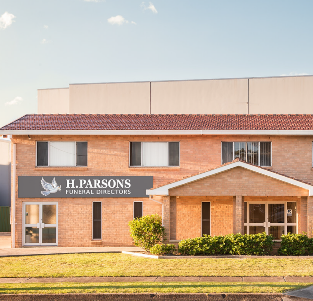 A brick building with a red tiled roof houses "H. Parsons Funeral Directors." The sign features a white dove. The building has large windows, a front garden, and a small covered entrance. The sky is clear and sunny. by H Parsons Funeral Directors