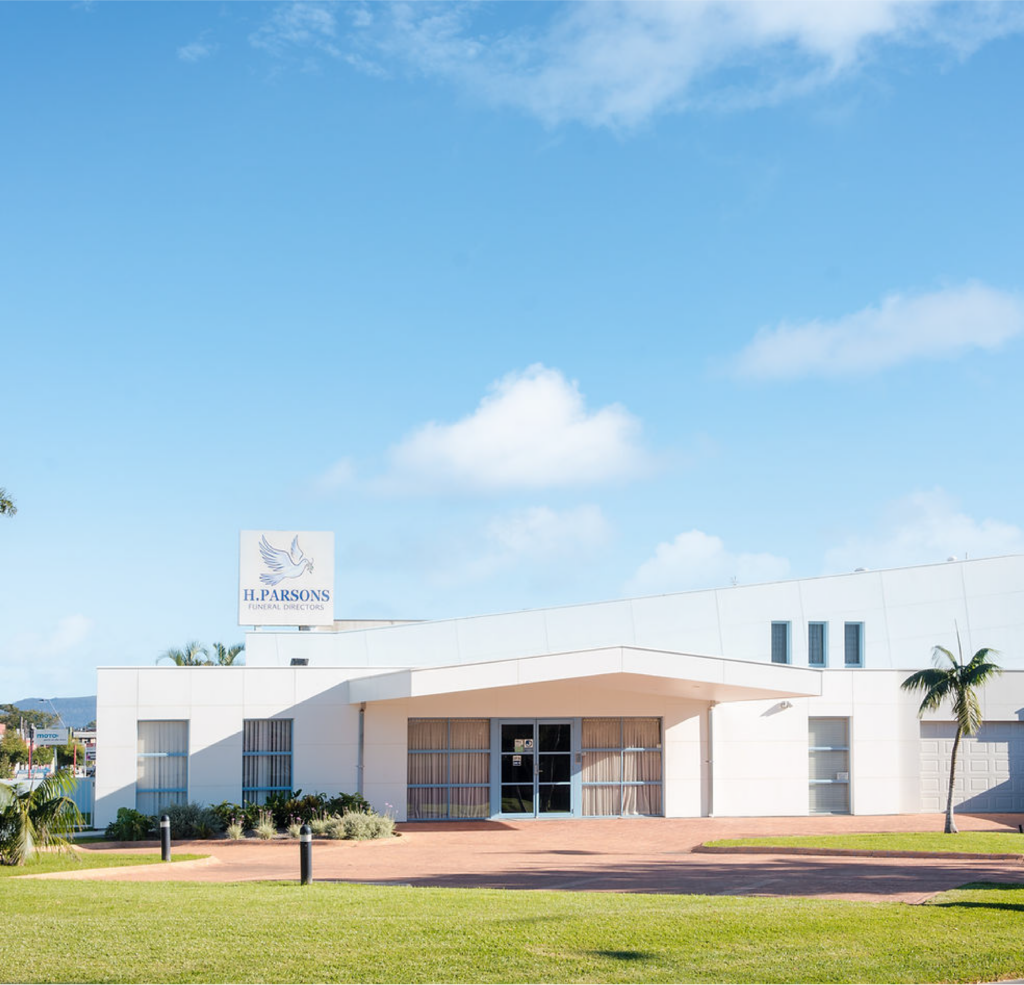 A modern white building with an H. Parsons sign and a dove logo above the entrance, surrounded by green grass, palm trees, and a clear blue sky. by H Parsons Funeral Directors