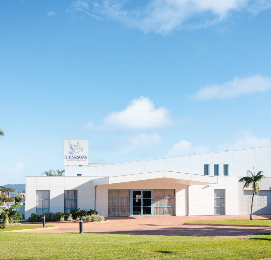 A modern white building with a flat roof and a sign reading "H. Parsons" featuring a dove logo; palm trees and green lawn surround the entrance under a clear blue sky. by H Parsons Funeral Directors