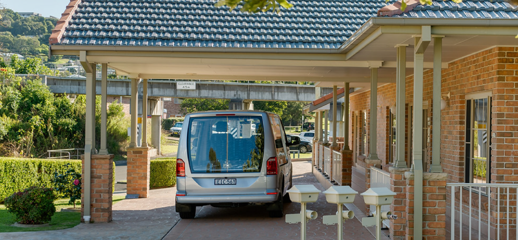A silver van is parked in the drive of a brick house with a tiled roof, covered carport, and manicured lawn. Three postboxes stand by the drive, and leafy trees and hedges surround the property. by H Parsons Funeral Directors