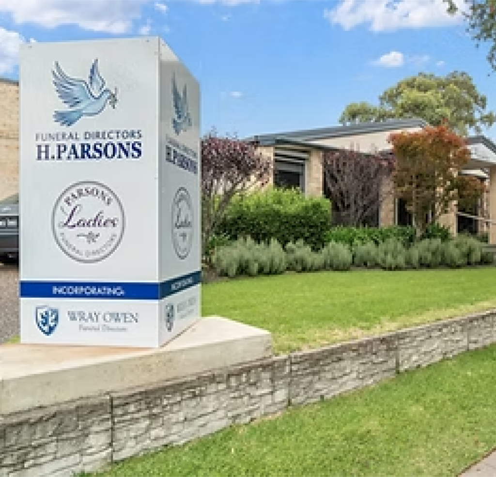 A large white sign for H. Parsons Funeral Directors stands on a lawn in front of a modern, single-storey building with greenery and trees. The sign features a dove and company logos. by H Parsons Funeral Directors