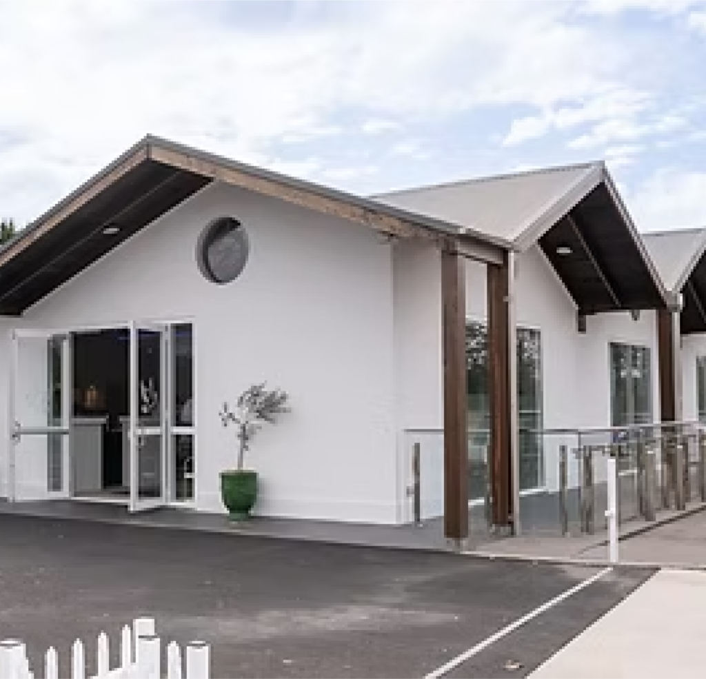 A modern white building with large glass doors, wooden beams, and a sloped roof, set on a paved area with a green plant by the entrance and a glass balustrade along one side. by H Parsons Funeral Directors