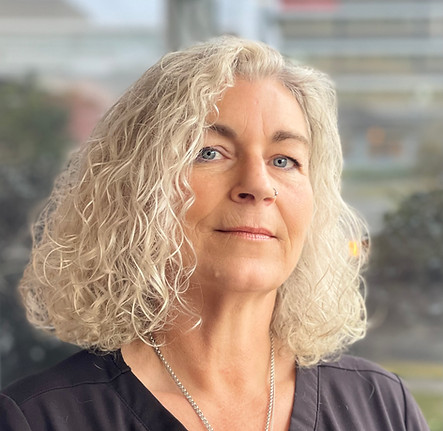 A woman with curly, shoulder-length grey hair and blue eyes wears a black top and a silver necklace, standing in front of a blurred background with natural light. by H Parsons Funeral Directors