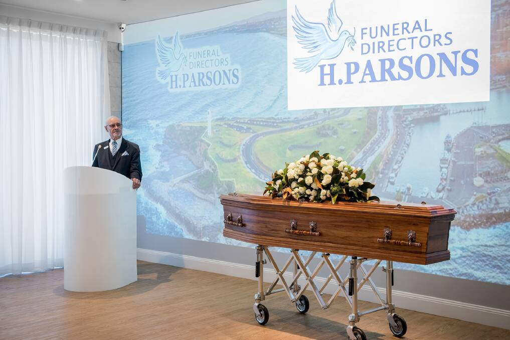 A man in a suit stands at a lectern next to a coffin adorned with flowers on a trolley. Behind him is a large screen displaying the H. Parsons Funeral Directors logo and an aerial view of a coastal area. by H Parsons Funeral Directors