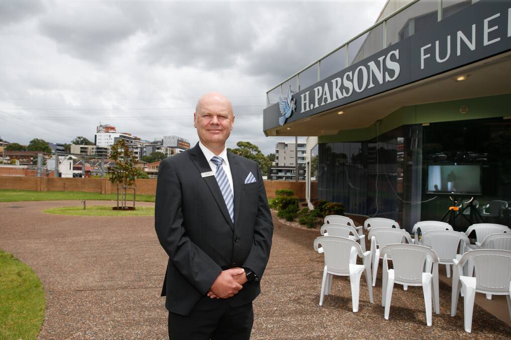 A man in a suit stands smiling outside H. Parsons Funeral Parlour. White plastic chairs are arranged near the entrance, with buildings and cloudy skies in the background. by H Parsons Funeral Directors