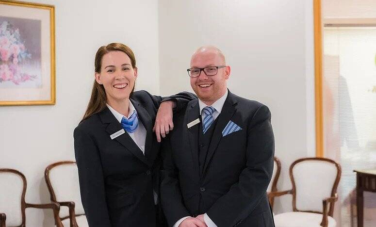 Two people in professional attire, both smiling, stand in a bright room with several empty chairs and a framed floral painting on the wall behind them. One person rests their arm on the other's shoulder. by H Parsons Funeral Directors