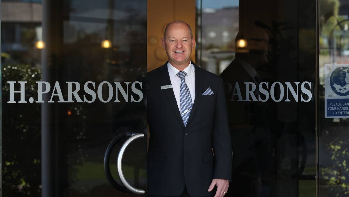 A man in a suit stands smiling at the entrance of a building with glass doors displaying the name "H. PARSONS." He wears a striped tie, name badge, and a pocket handkerchief. The background shows reflections of the outside. by H Parsons Funeral Directors