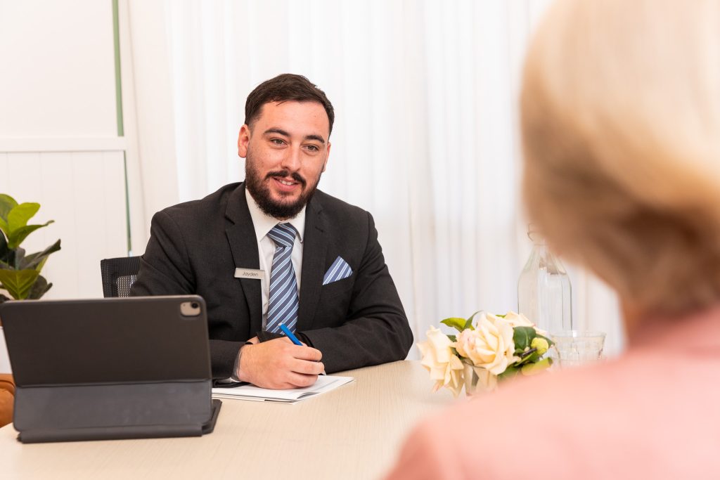 A man in a suit sits at a desk with a tablet, paper, and pen, speaking to a person whose back is to the camera. Flowers and a glass of water adorn the table, creating a professional yet friendly setting for legal services. by H Parsons Funeral Directors