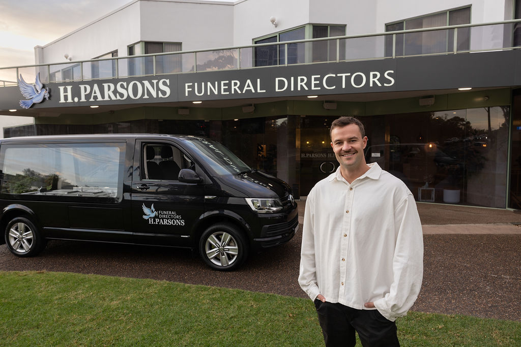 A smiling man in a white shirt stands in front of the H Parsons Funeral Directors building, with a black H Parsons funeral vehicle parked outside. by H Parsons Funeral Directors