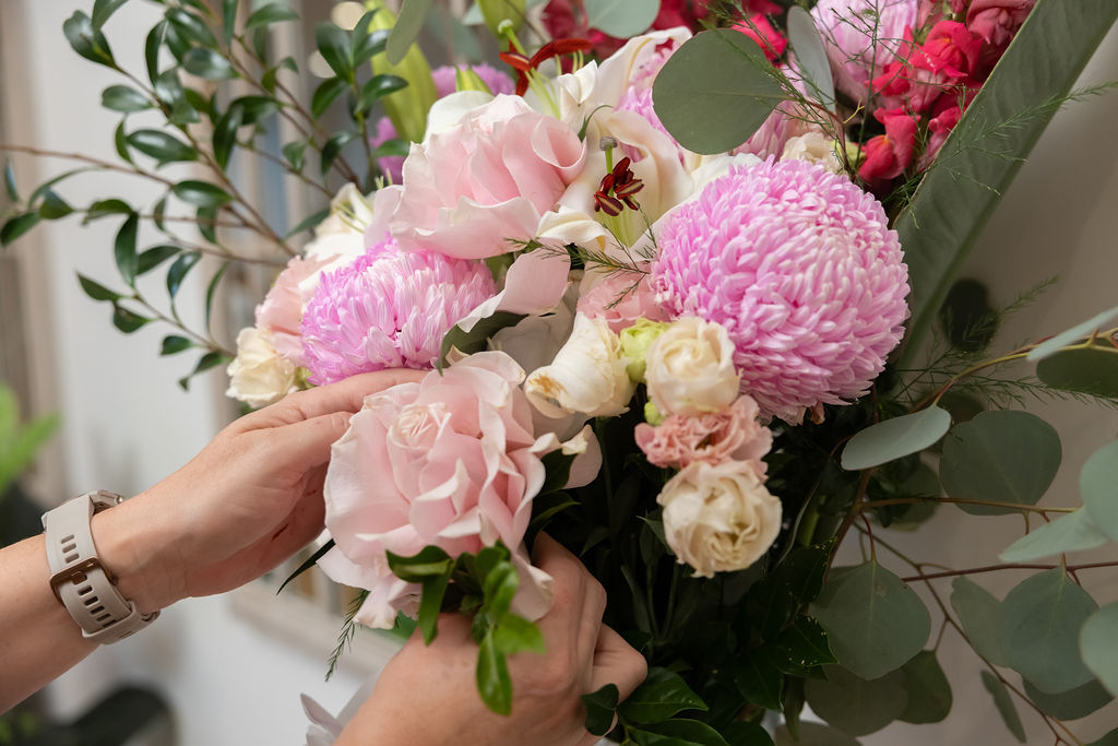 Hands arranging a bouquet of flowers with pink chrysanthemums, light pink roses, white blooms, and green foliage—a comforting gesture often seen when someone dies abroad. The person is wearing a beige smart watch on their left wrist. by H Parsons Funeral Directors